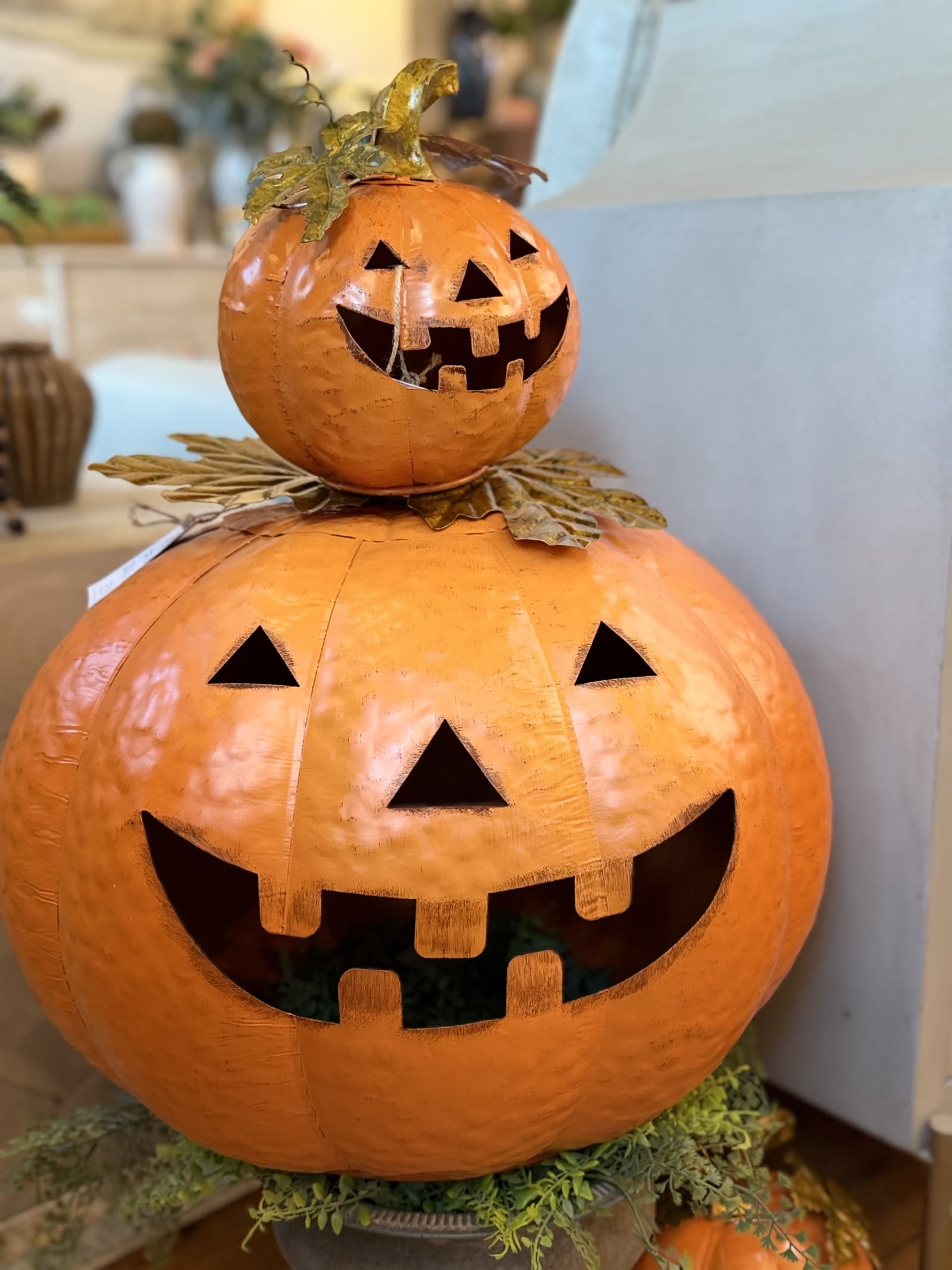 A pair of stacked orange metal jack-o-lantern lanterns on a greenery filled urn inside the MONA store