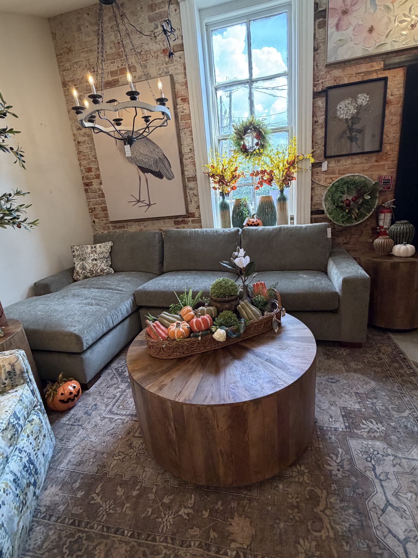 A grey sectional sofa and round mango wood coffee table styled with a basket of gourds under a wood bead chandelier at MONA of Waxhaw